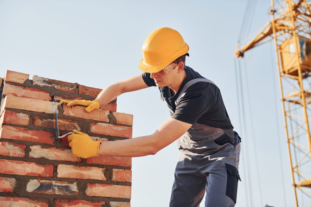 A man wearing overalls and a hard hat is diligently building a brick chimney, showcasing his craftsmanship on the wall.