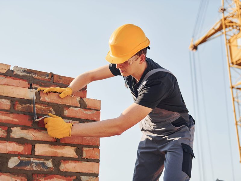 A man wearing overalls and a hard hat is diligently building a brick chimney, showcasing his craftsmanship on the wall.