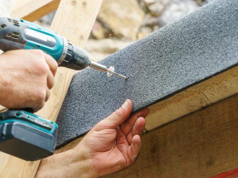 A man operates a drill to install roofing material, ensuring proper placement for effective gutter service.